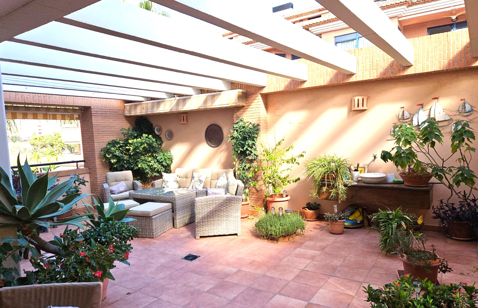 Amplia terraza privada con suelo de terracota y vigas de pérgola blancas. Incluye mobiliario de mimbre, paredes de ladrillo visto y variada vegetación mediterránea.