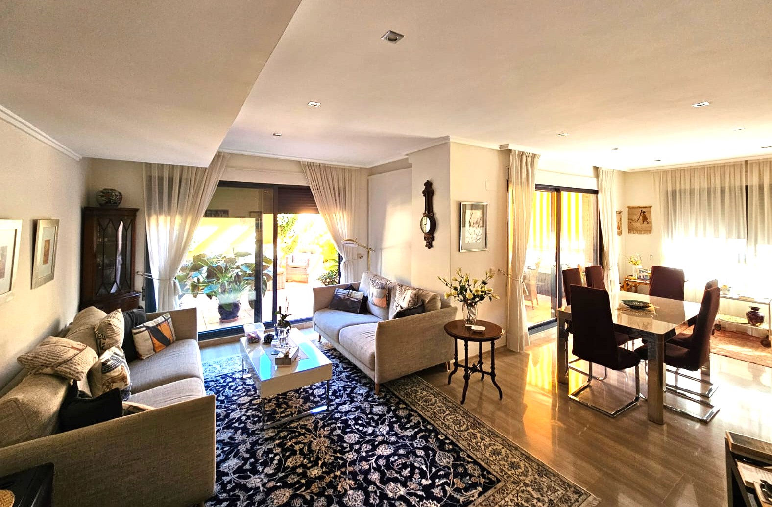 Amplio salón-comedor con suelos de piedra pulida, iluminación empotrada y grandes ventanales correderos que conectan con una terraza exterior ajardinada.