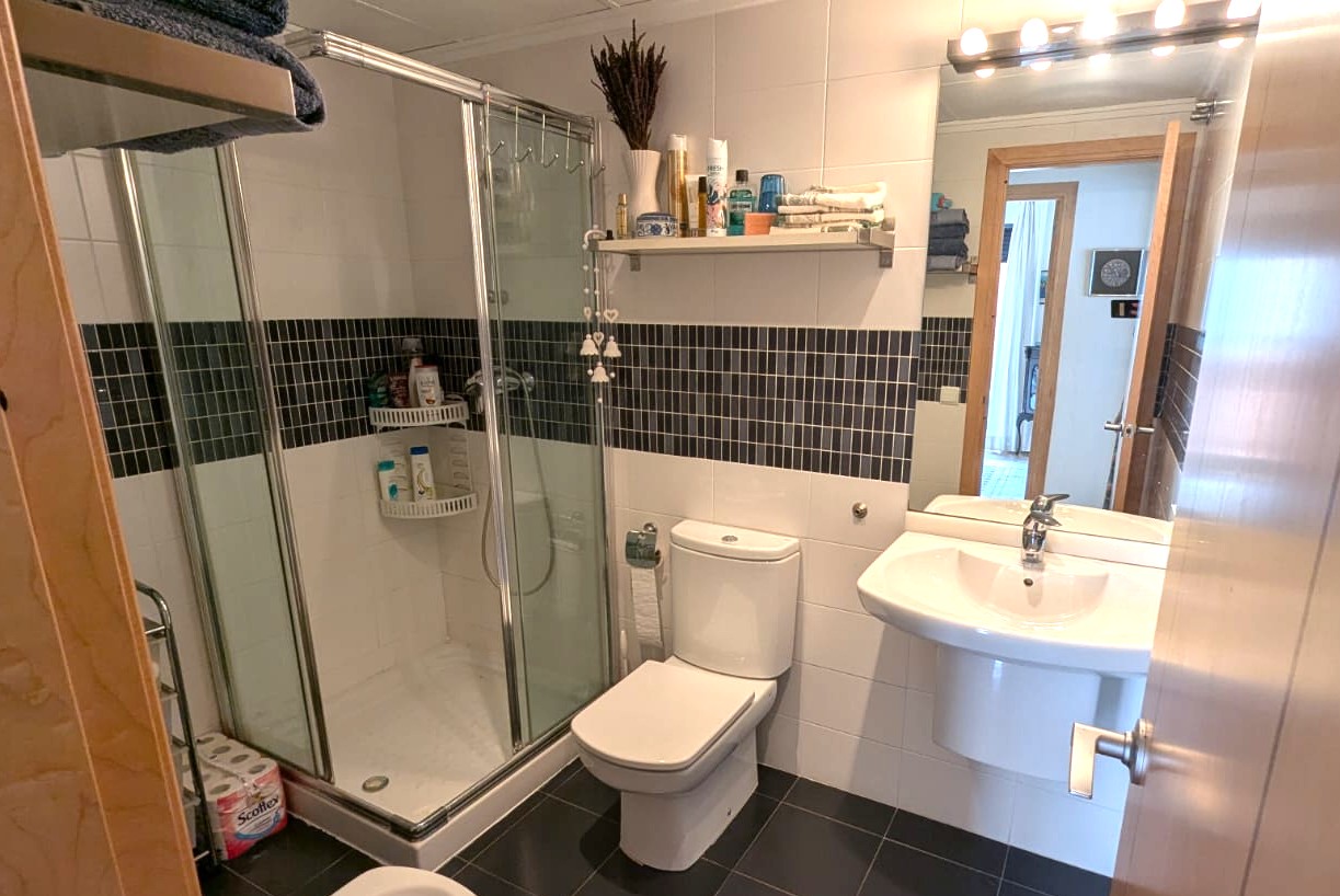 Baño equipado con ducha de cabina acristalada, revestimiento cerámico blanco con detalle de mosaico oscuro y suelo de gres en tono antracita.