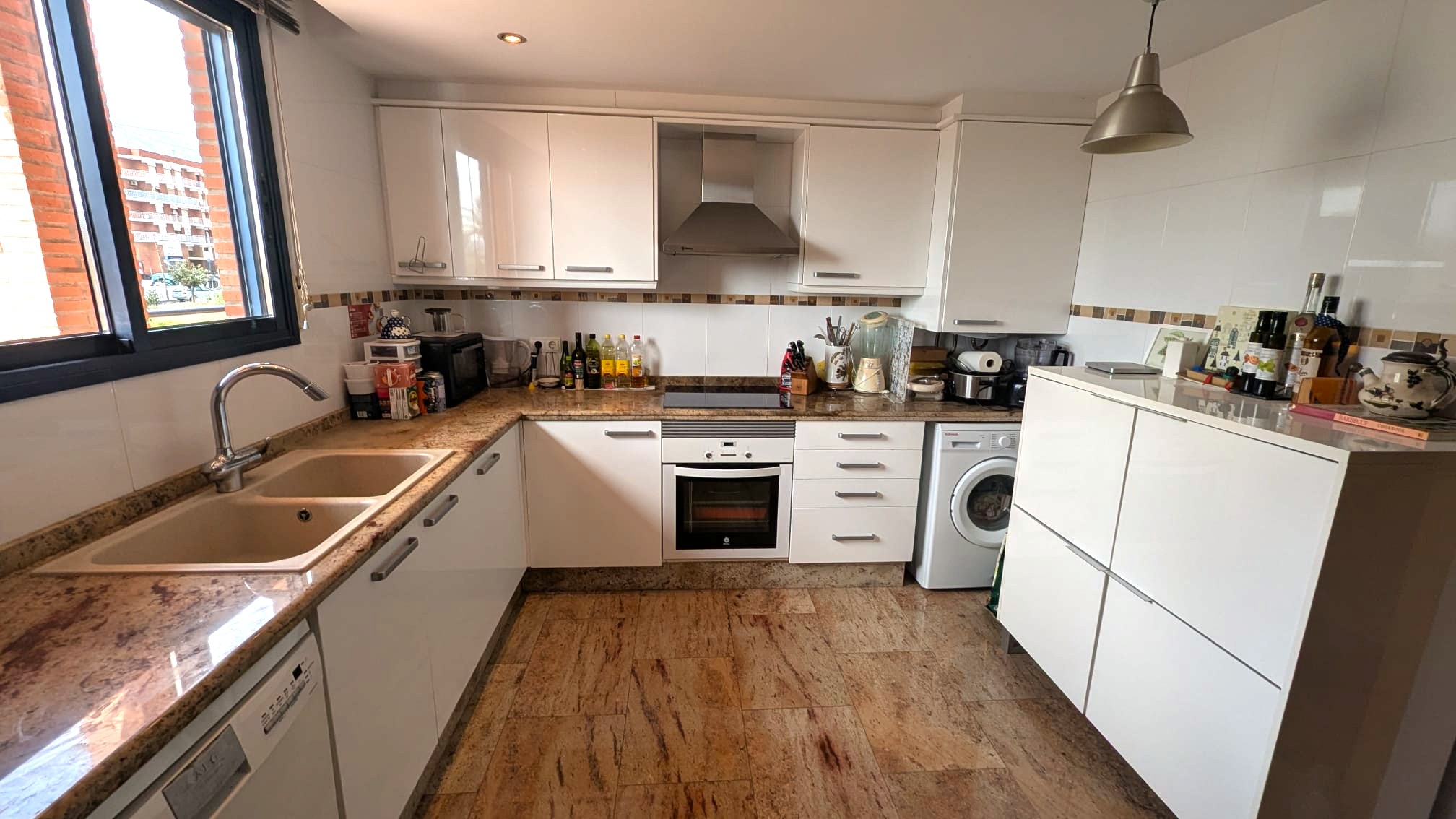 Cocina funcional con armarios blancos, encimeras de granito y suelos de piedra natural. Incluye electrodomésticos integrados y una ventana con carpintería oscura.