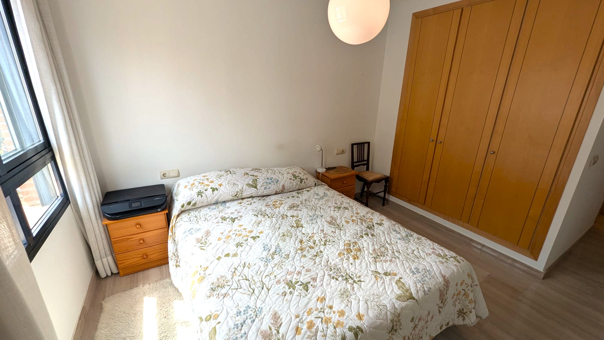 Dormitorio con armario empotrado de madera de cuatro puertas, cama doble y mesitas de noche a juego. Dispone de suelo de tarima clara y ventana con carpintería negra.