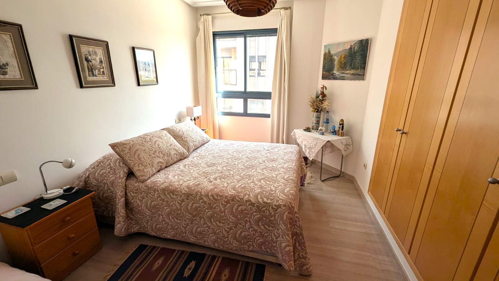 Dormitorio con armarios empotrados de madera, suelos de tarima clara y ventana con luz natural. Dispone de mobiliario funcional y decoración clásica en tonos cálidos.