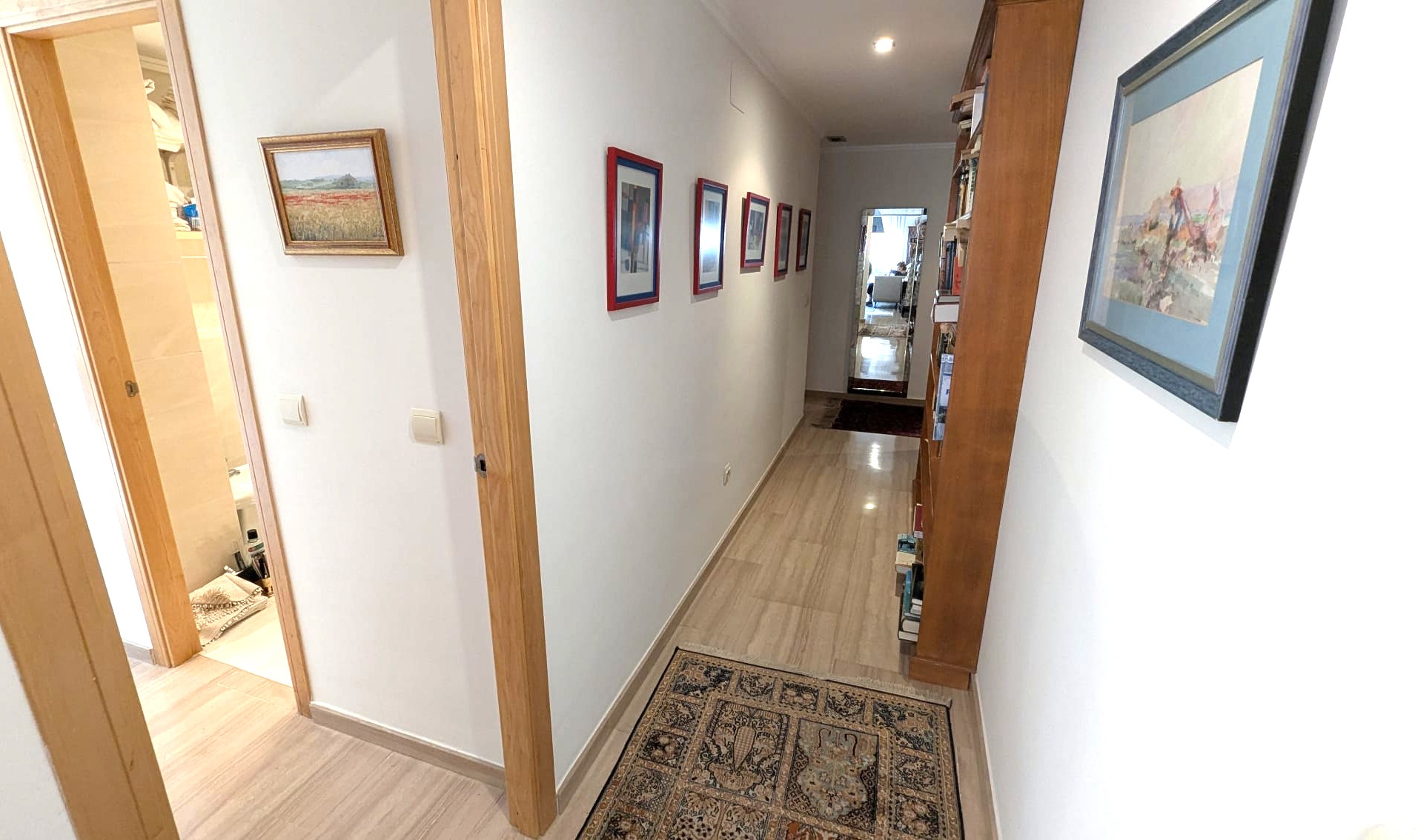 Pasillo luminoso con suelos de piedra pulida, marcos de puertas en madera y una estantería integrada. Incluye iluminación empotrada y cuadros decorativos.