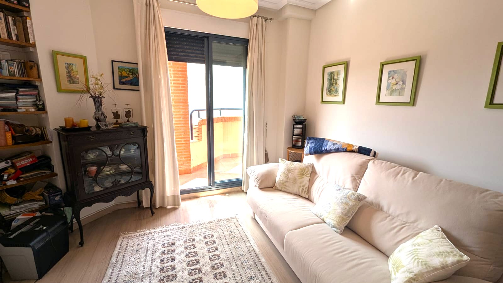 Salón luminoso con sofá beige, vitrina clásica de madera oscura y acceso directo a terraza privada mediante puertas correderas de cristal.