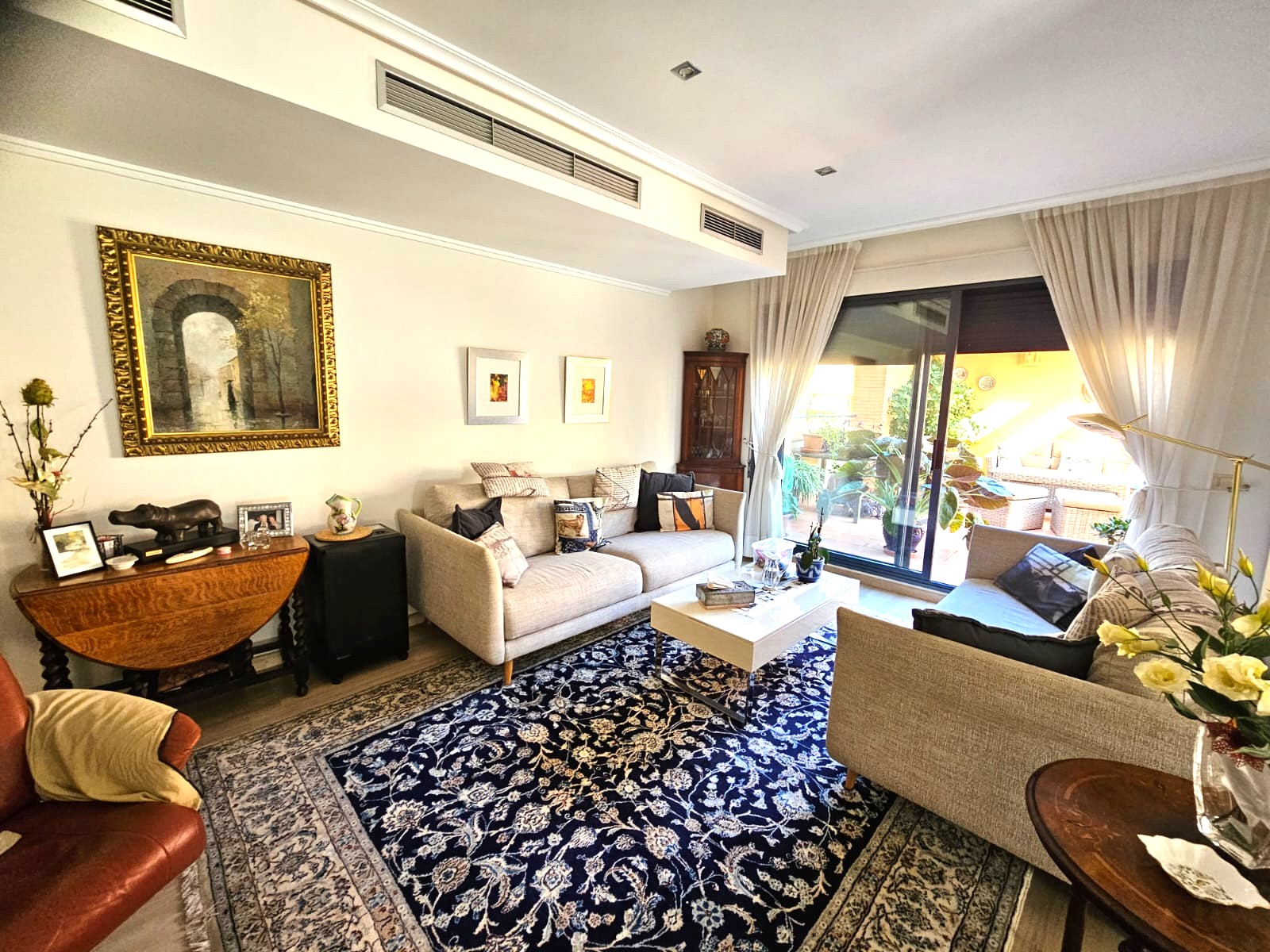 Salón luminoso con sofás de tela, alfombra de diseño clásico y acceso a terraza privada. Incluye sistema de climatización por conductos y techos con iluminación empotrada.