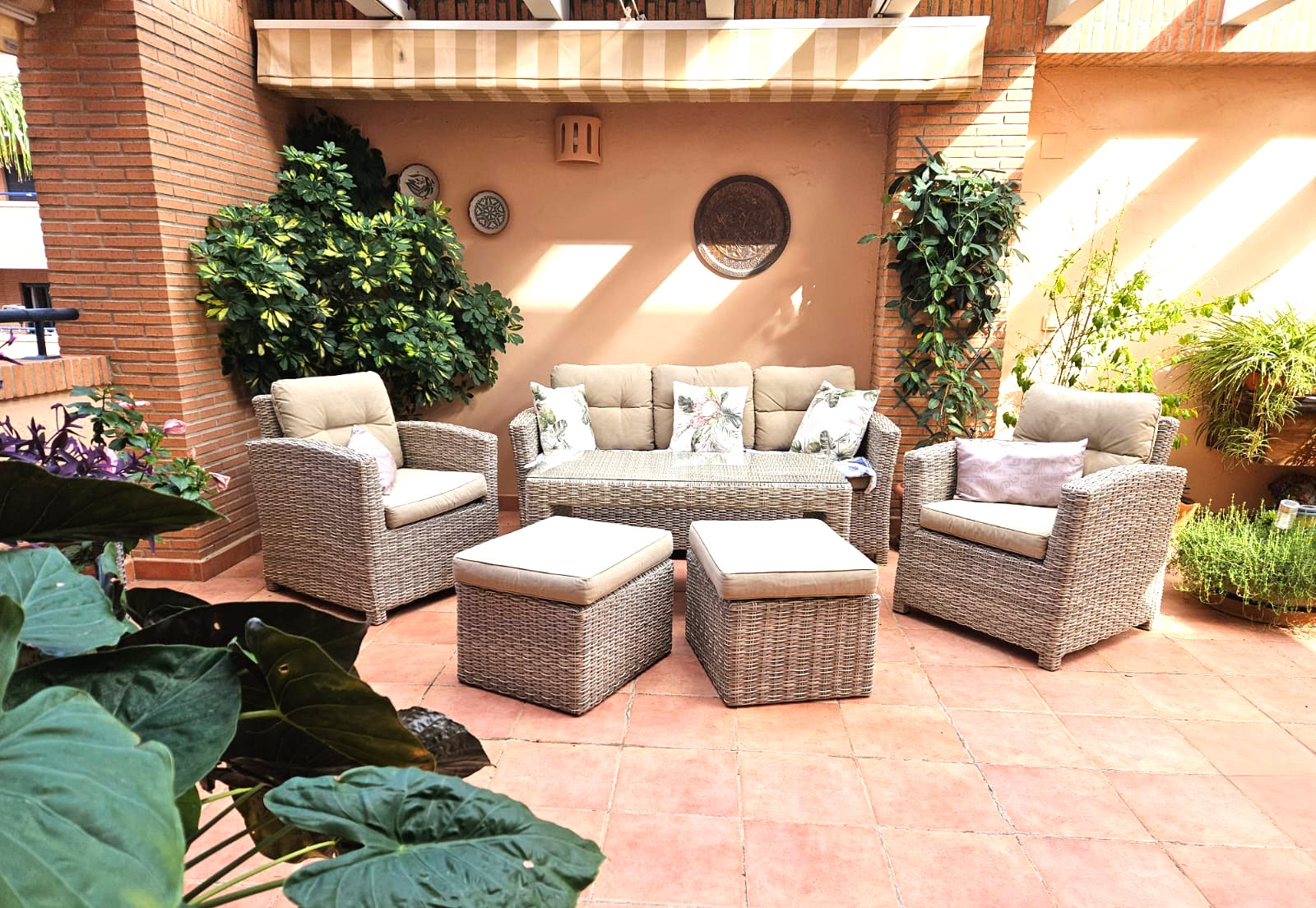 Terraza privada con suelo de terracota, mobiliario de ratán sintético y zona de estar bajo toldo extensible, rodeada de plantas y paredes de ladrillo visto.