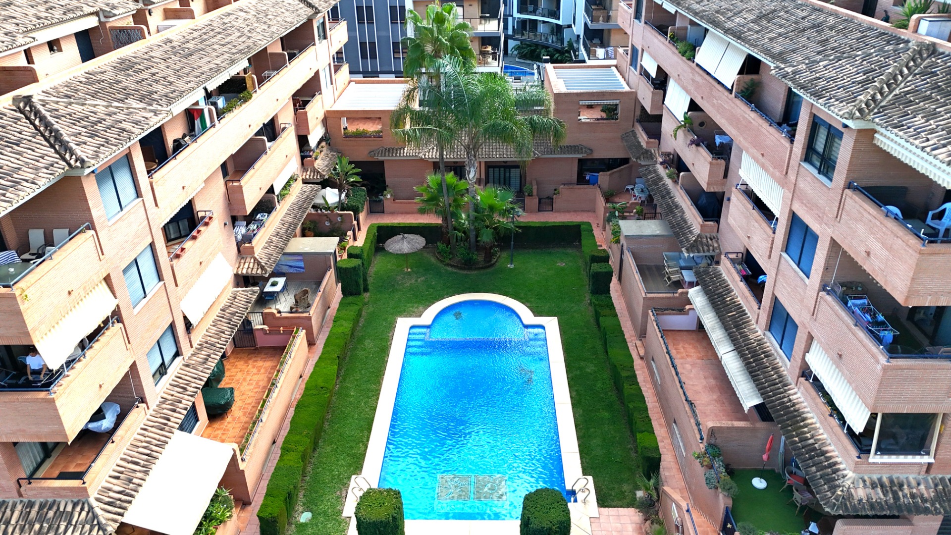 Vista elevada de la zona comunitaria con piscina azul, césped cuidado y palmeras, rodeada por edificios de ladrillo con terrazas privadas en Jávea.