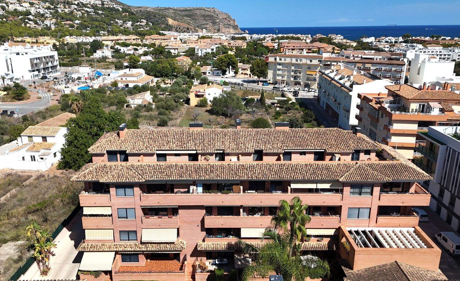 Vista elevada de un complejo de apartamentos de ladrillo con tejados de terracota, balcones con toldos y vistas panorámicas hacia el mar Mediterráneo en Jávea.
