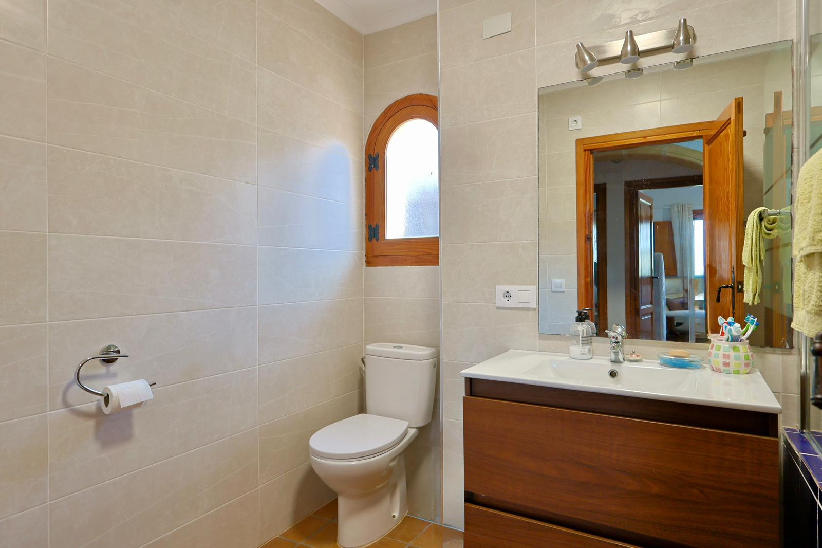 Baño con mueble de madera oscura, lavabo integrado y una característica ventana de madera en arco sobre azulejos beige y suelos de gres cálidos.