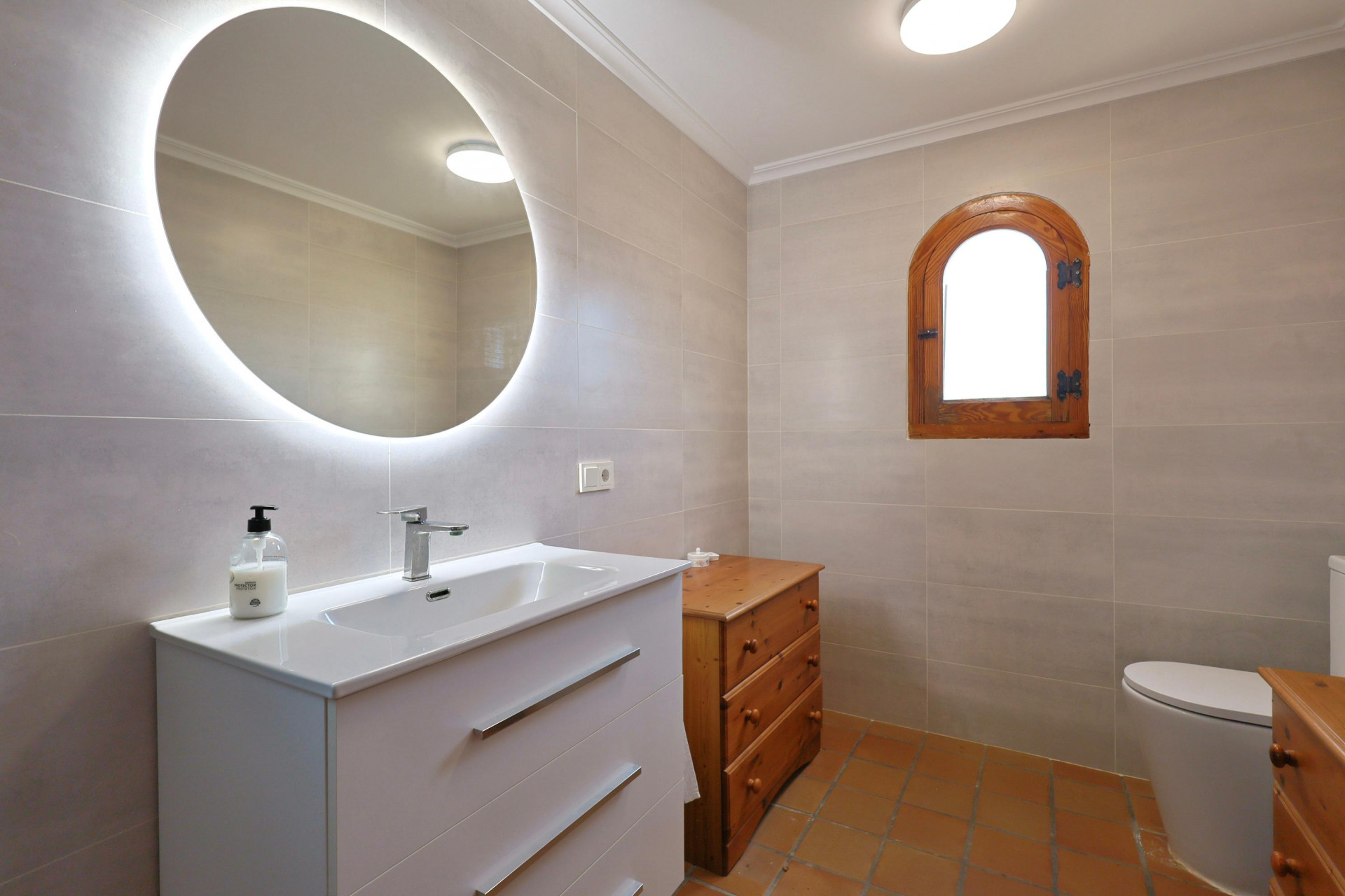 Baño moderno con espejo circular retroiluminado, mueble de lavabo blanco, suelo de terracota y una ventana de madera con arco tradicional.
