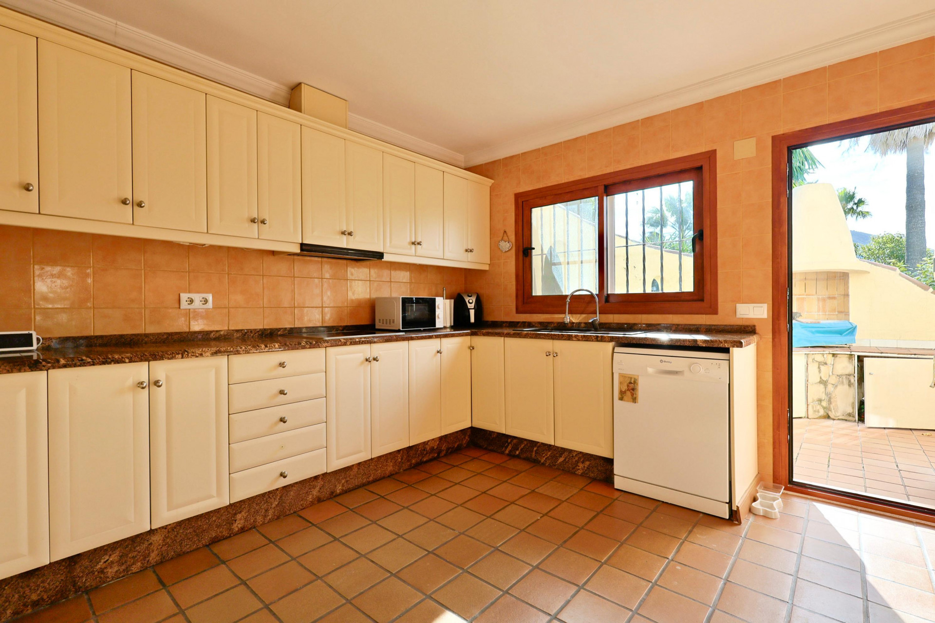 Cocina equipada con armarios color crema, encimera de granito y suelos de terracota. Dispone de acceso directo al exterior y ventana de madera sobre el fregadero.
