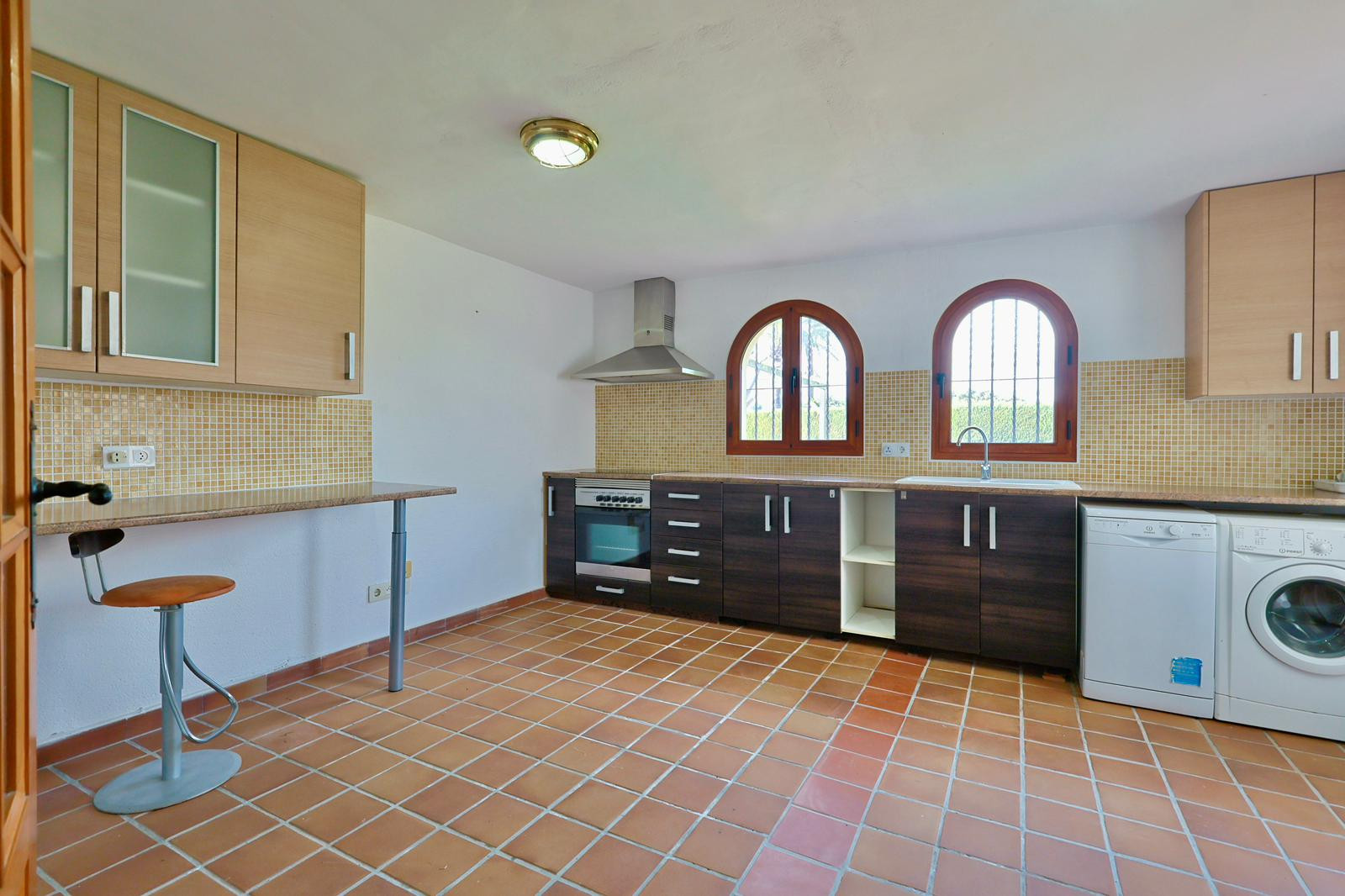Cocina funcional con suelos de terracota, armarios bitono, ventanas de arco de madera y electrodomésticos integrados. Incluye zona de desayuno y salpicadero de mosaico beige.
