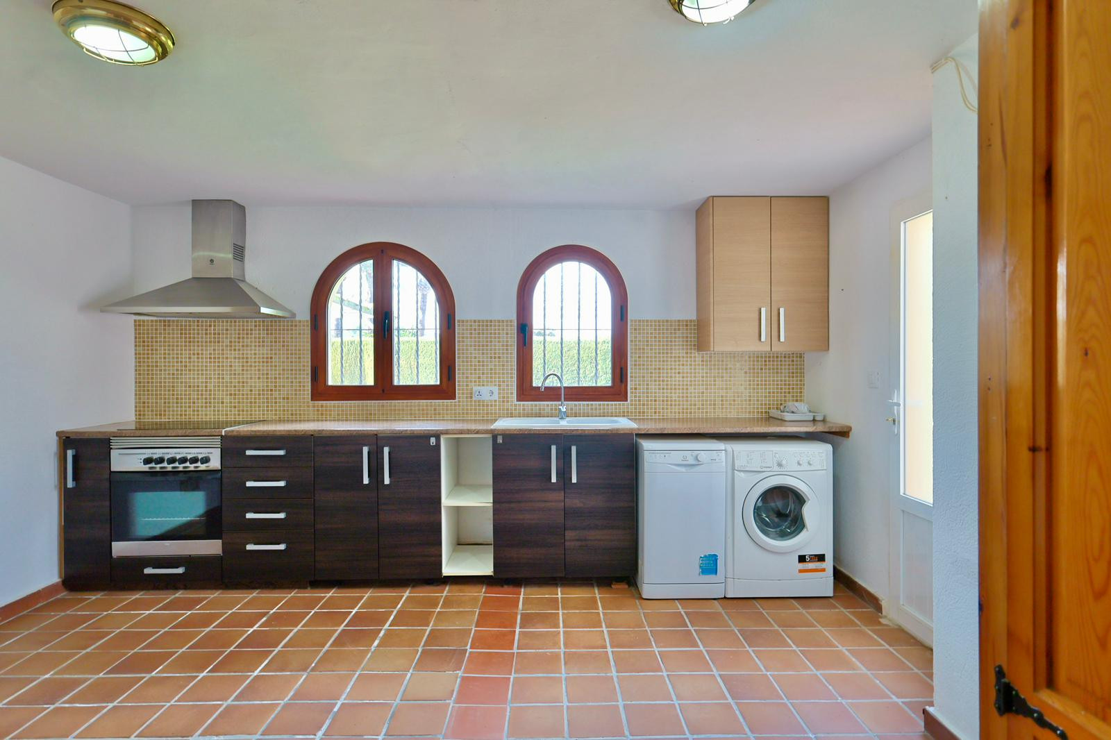 Cocina funcional con zona de lavandería, armarios oscuros, suelo de terracota y ventanas de arco. Incluye electrodomésticos integrados y salpicadero de mosaico.
