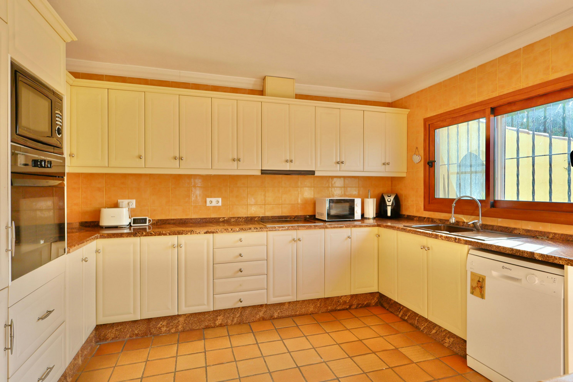 Cocina tradicional con armarios color crema, encimeras de granito marrón y azulejos de terracota. Incluye electrodomésticos integrados y una ventana con marco de madera.