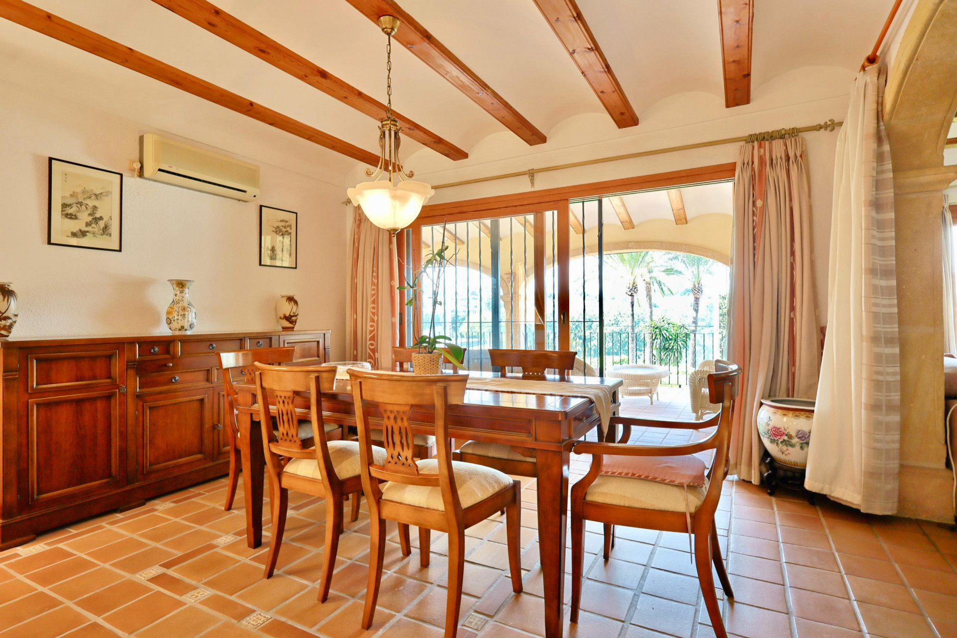 Comedor con vigas de madera vistas, suelo de terracota y aparador clásico. Grandes puertas correderas de cristal dan acceso a una naya con vistas exteriores.