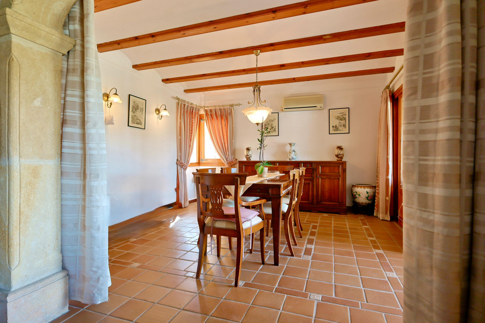 Comedor de estilo rústico con vigas de madera, suelos de barro cocido y arco de piedra tosca. Dispone de mobiliario de madera maciza y luz natural.