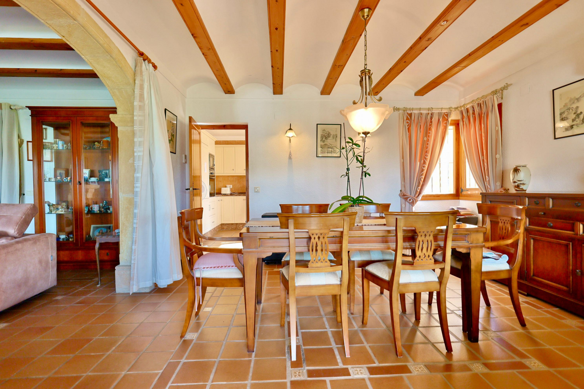 Comedor rústico con vigas de madera, suelos de terracota y un arco de piedra tosca. Incluye mesa de madera maciza y acceso visual a la cocina independiente.