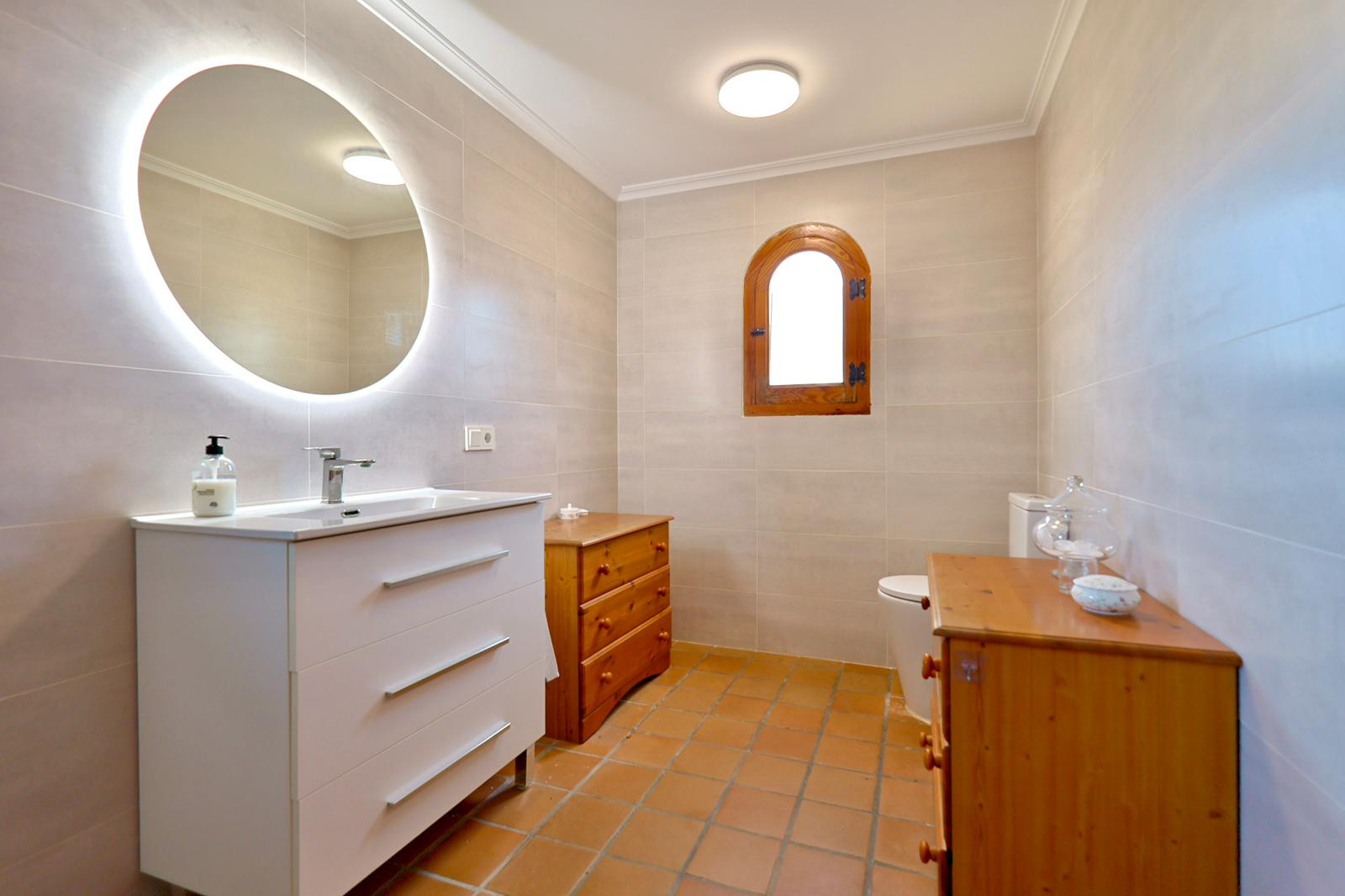 Cuarto de baño con lavabo blanco, espejo circular retroiluminado y suelos de terracota. Incluye una ventana de arco en madera y mobiliario auxiliar de pino.