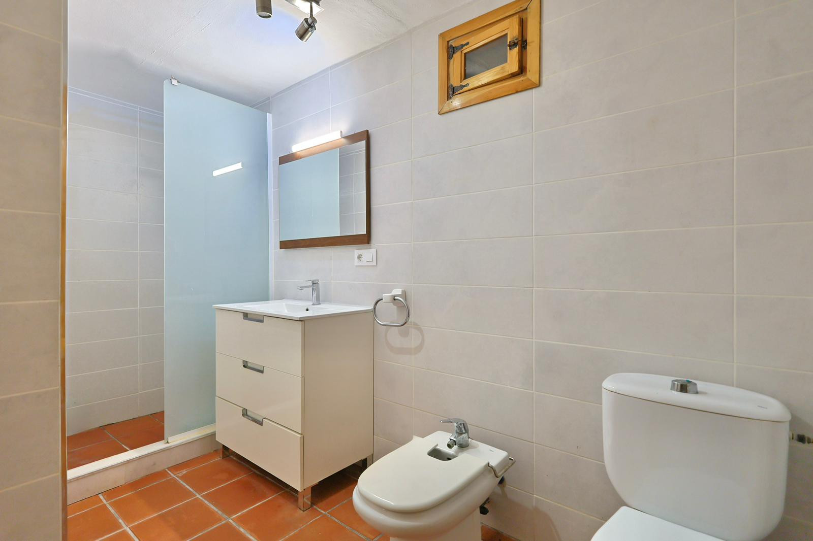 Cuarto de baño con suelo de terracota, mueble de lavabo blanco de tres cajones, bidé y ducha con mampara de cristal translúcido.