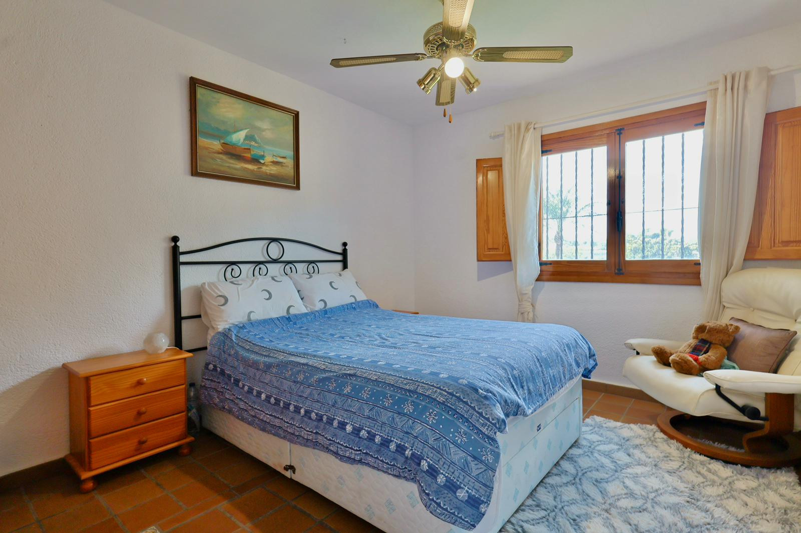 Dormitorio con cabecero de forja, suelo de baldosa de barro cocido y ventana de madera con contraventanas. Dispone de ventilador de techo y luz natural.