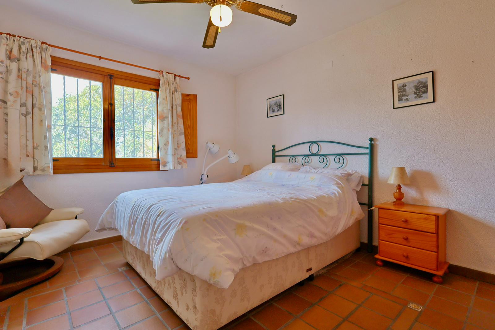 Dormitorio con suelo de baldosas de barro, ventana de madera con rejas de seguridad, cabecero de forja verde y ventilador de techo. Incluye mesita de noche de madera.