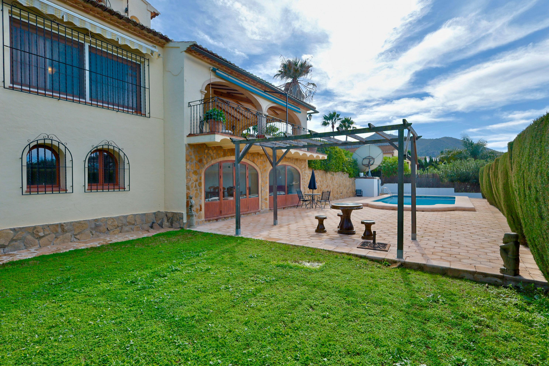 Exterior de la villa con fachada inferior de piedra, puertas arqueadas, pérgola de madera sobre terraza de gres y piscina privada junto al césped.
