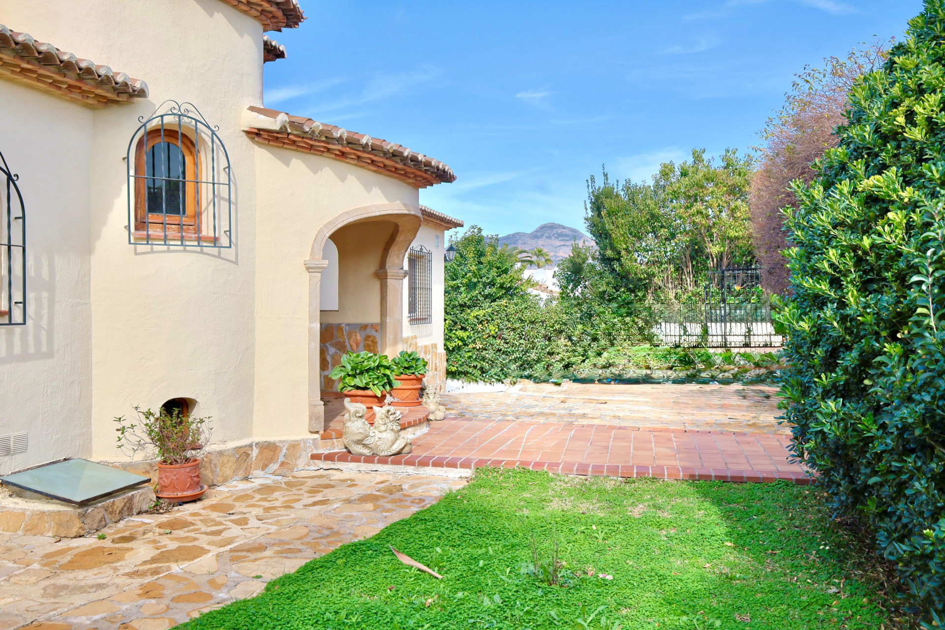 Exterior de villa mediterránea con porche de piedra, suelos de terracota y ventanas de madera con rejas de hierro forjado bajo el cielo despejado.