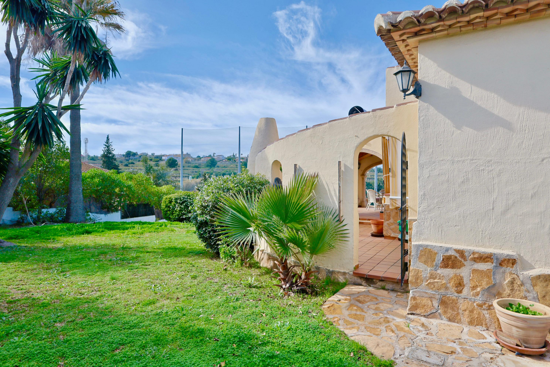 Fachada de la villa con base de piedra natural, jardín con palmeras y acceso arqueado a la terraza de terracota con vistas al entorno del club de golf.