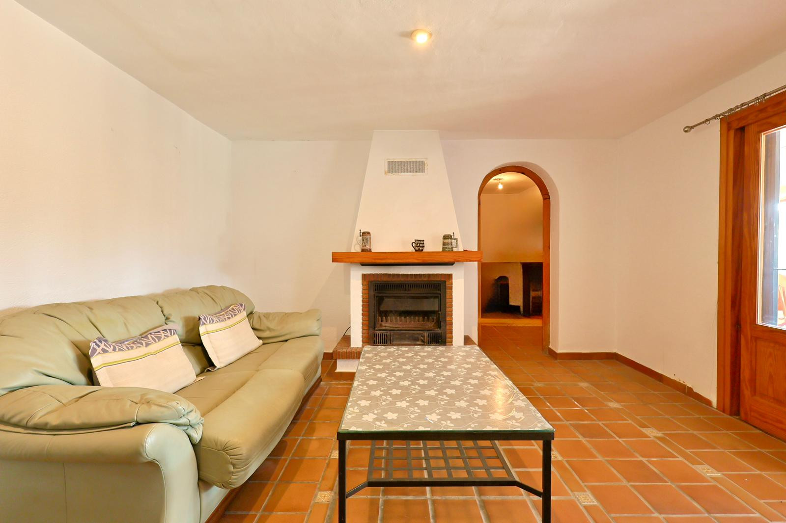 Salón con chimenea de obra y repisa de madera, suelos de baldosa de terracota y un arco que conecta con las estancias contiguas.