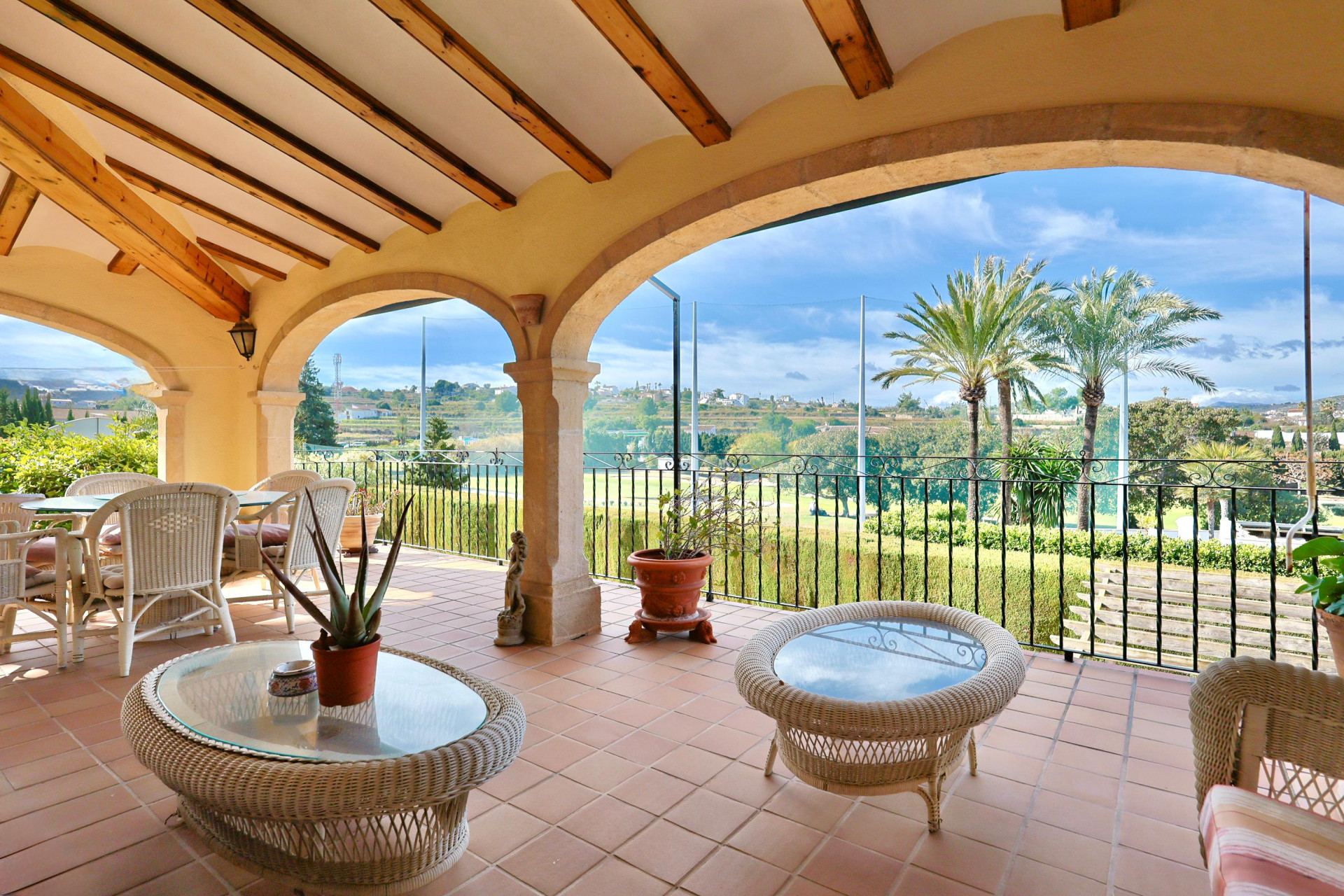 Terraza cubierta con arcos de piedra tosca, vigas de madera a la vista y suelo de barro, con vistas despejadas al campo de golf y palmeras.