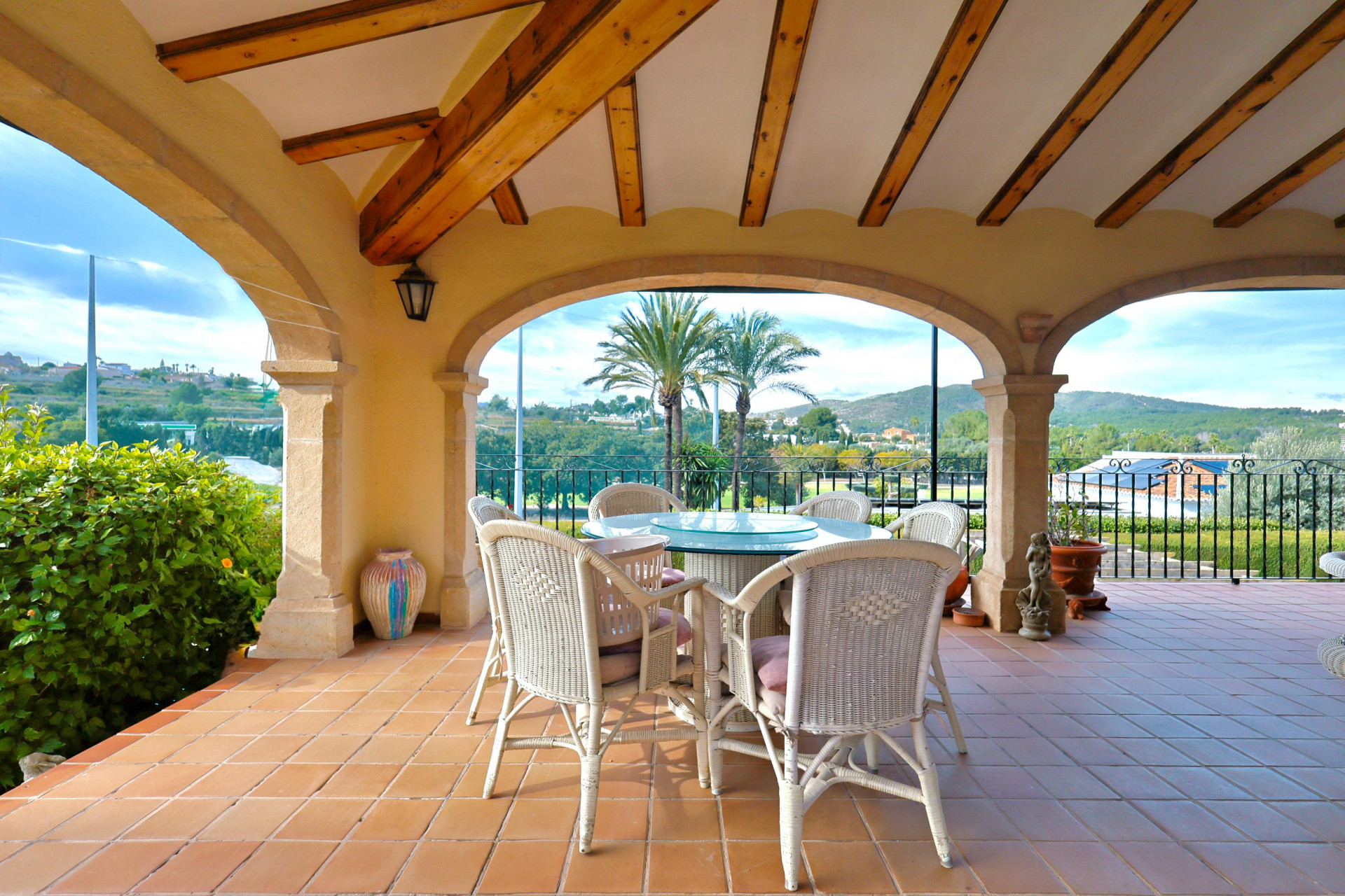 Terraza cubierta con arcos de piedra Tosca y vigas de madera. Incluye comedor exterior con muebles de mimbre y vistas despejadas al paisaje mediterráneo y palmeras.