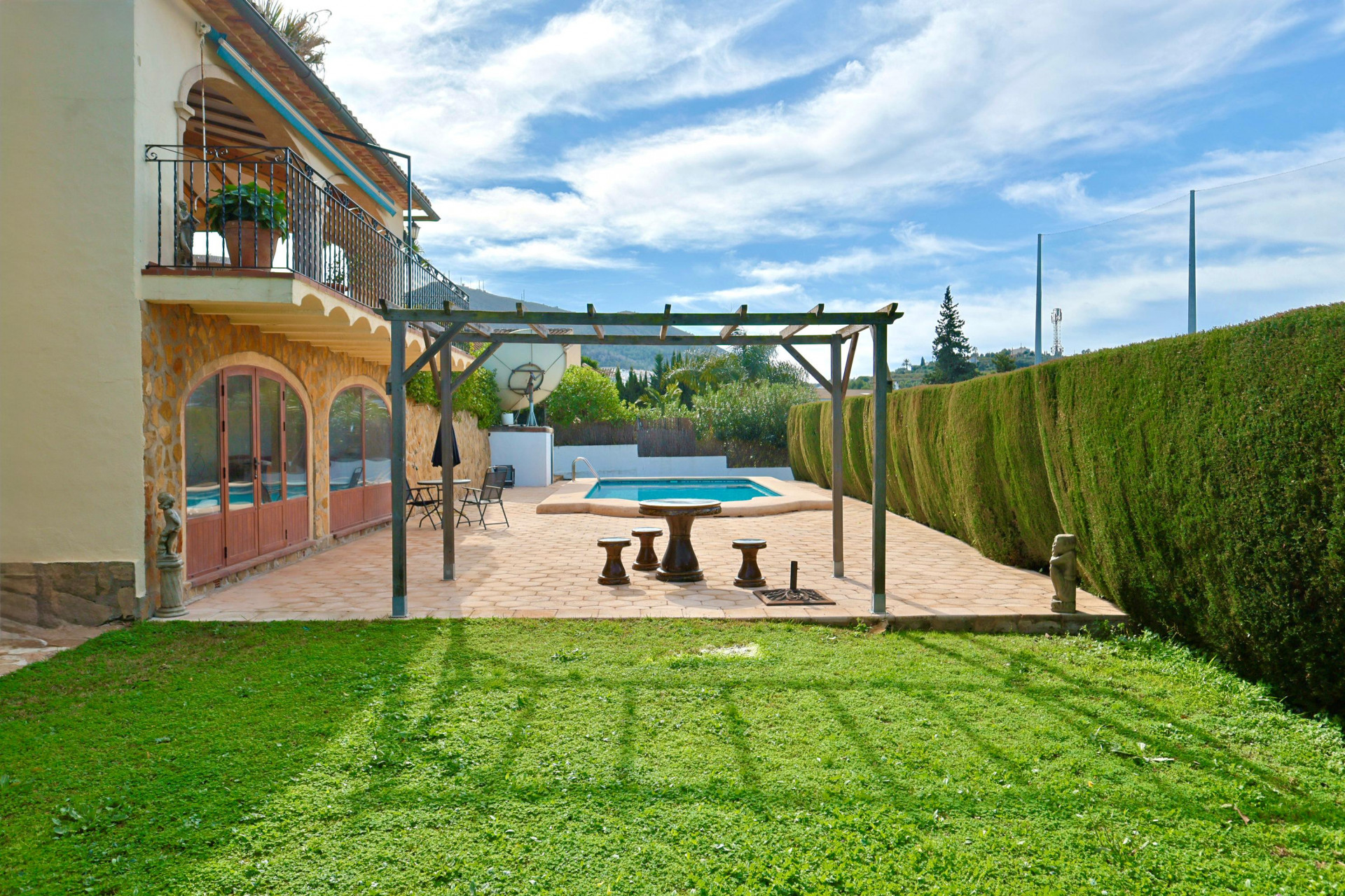 Terraza exterior con pérgola de madera y asientos de piedra, junto a una piscina privada y jardín, destacando la fachada de piedra con ventanales en arco.