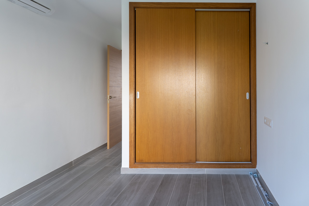 Dormitorio con armario empotrado de puertas correderas en madera natural, suelos de gres con acabado efecto madera gris y climatización tipo split.