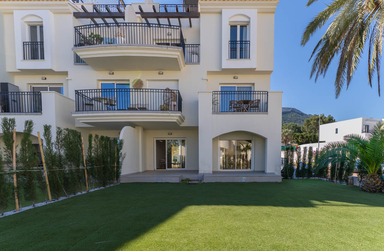 Fachada posterior de la planta baja con jardín privado de césped artificial y setos perimetrales, integrada en una urbanización mediterránea con vistas al Montgó.