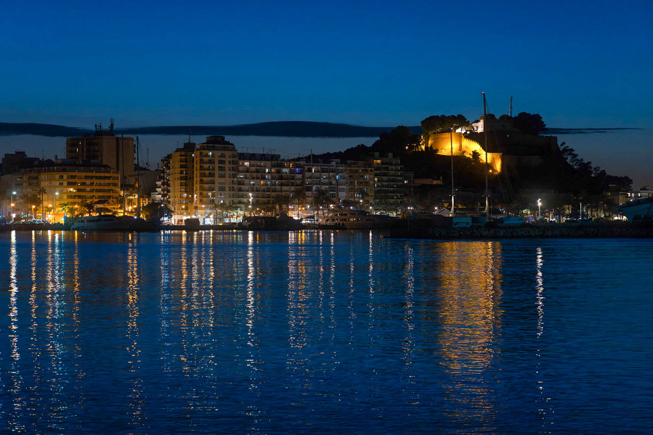 Vista nocturna del puerto de Dénia con el histórico castillo iluminado sobre la colina y los reflejos de las luces urbanas sobre las aguas del Mediterráneo.