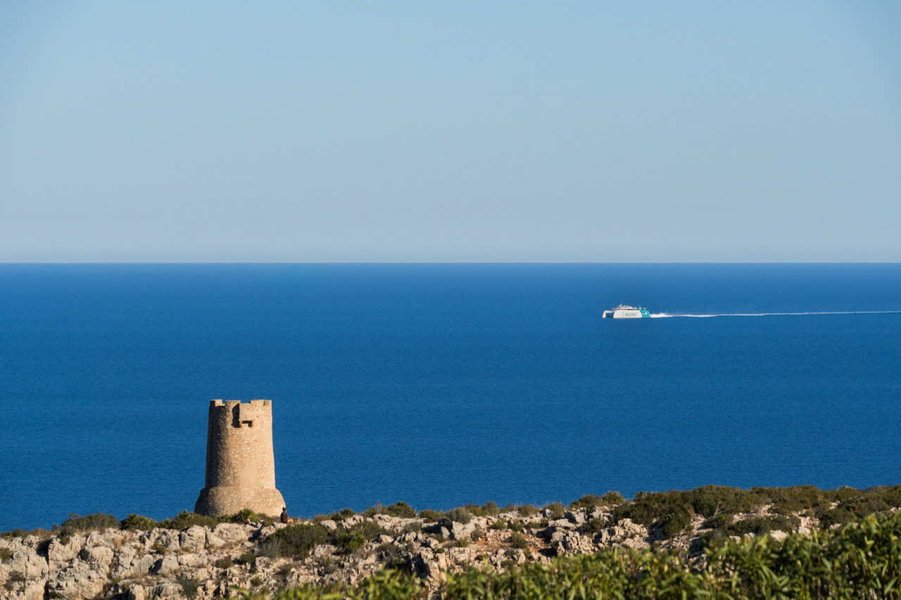 Vista panorámica de la Torre del Gerro sobre los acantilados de Dénia, con el mar Mediterráneo y un ferry de Baleària en el horizonte bajo un cielo despejado.