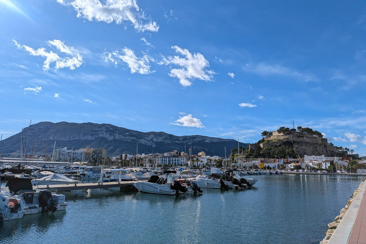 Vista panorámica del puerto deportivo de Dénia con embarcaciones de recreo, el castillo histórico sobre la colina y el macizo del Montgó al fondo bajo un cielo despejado.