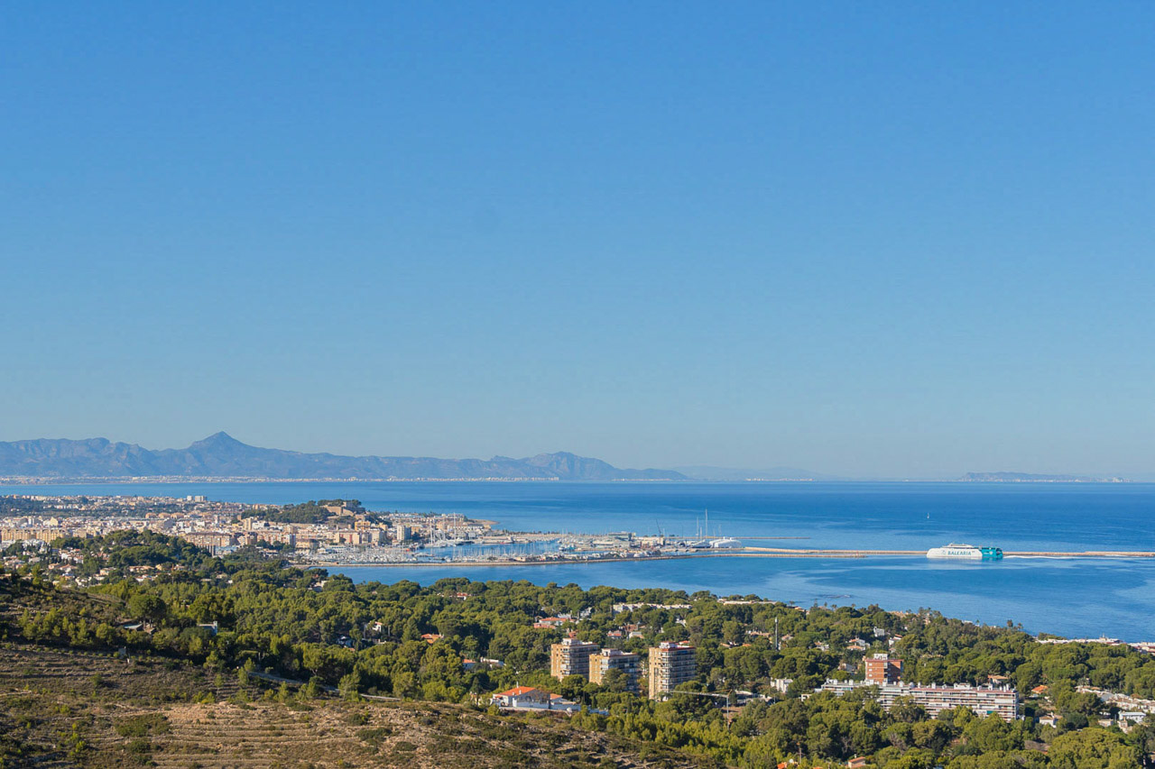 Vista panorámica elevada de Dénia, destacando el puerto deportivo, el castillo y el litoral mediterráneo bajo un cielo despejado con montañas al fondo.
