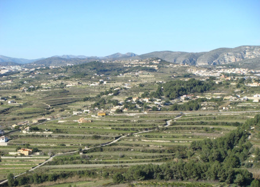 Vista panorámica de terrenos abancalados en Benitachell, destacando la topografía escalonada y la vegetación mediterránea con montañas al fondo.