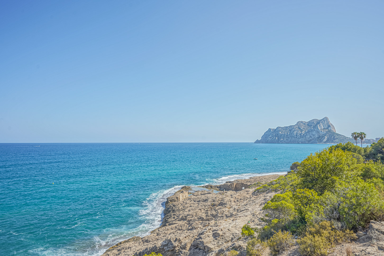 Vista panorámica de la costa mediterránea con el Peñón de Ifach al fondo, rodeada de vegetación autóctona y acantilados rocosos bajo un cielo despejado.