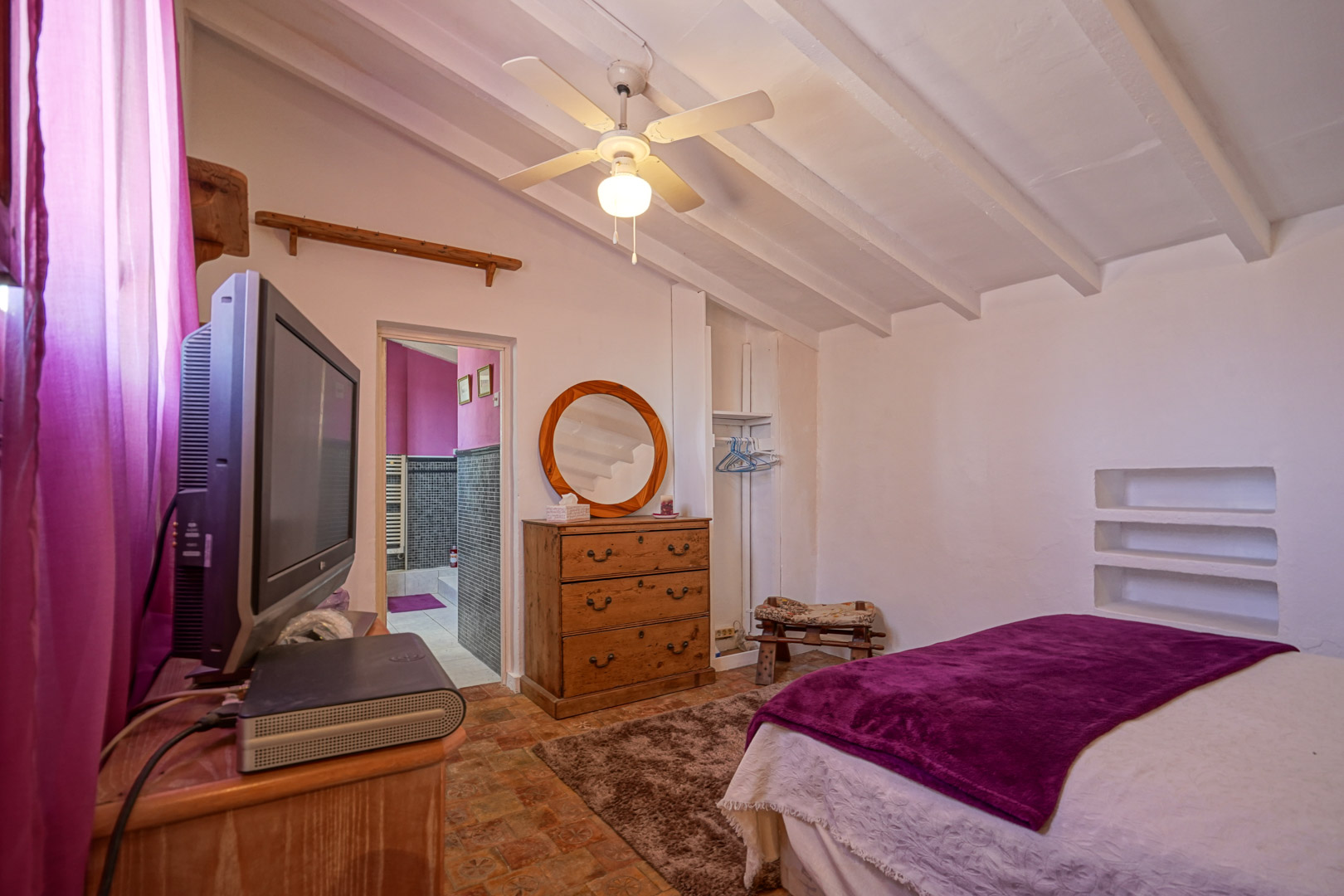 Dormitorio con techos inclinados de vigas blancas, ventilador de techo y acceso directo a un baño en suite con azulejos oscuros.