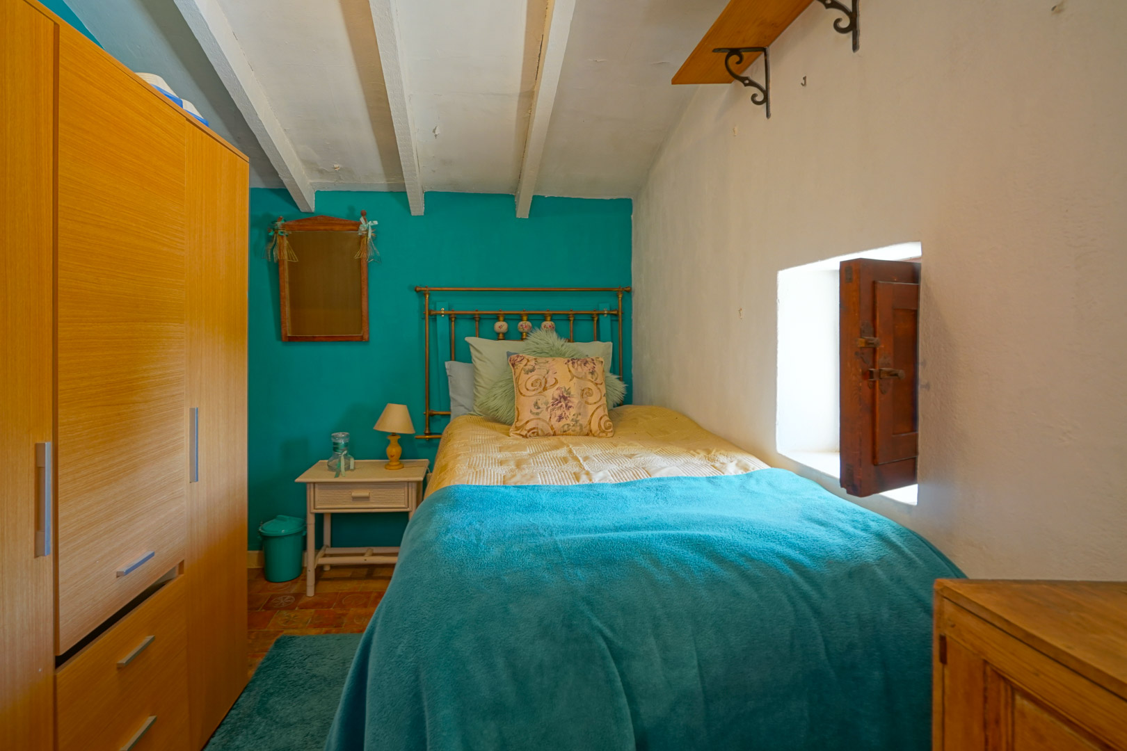 Dormitorio individual con vigas de madera blancas, paredes en contraste turquesa y suelos de terracota. Incluye un armario de madera y una ventana tradicional con contraventana.