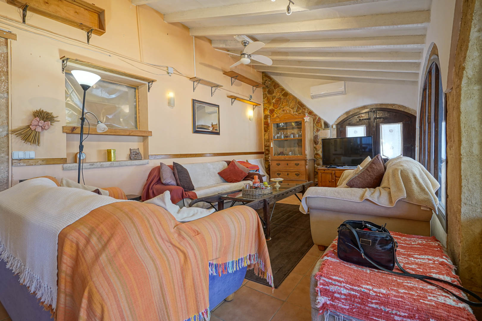 Salón con techos de vigas blancas inclinadas, paredes de piedra natural y arcos de madera tradicionales, equipado con aire acondicionado y ventilador.