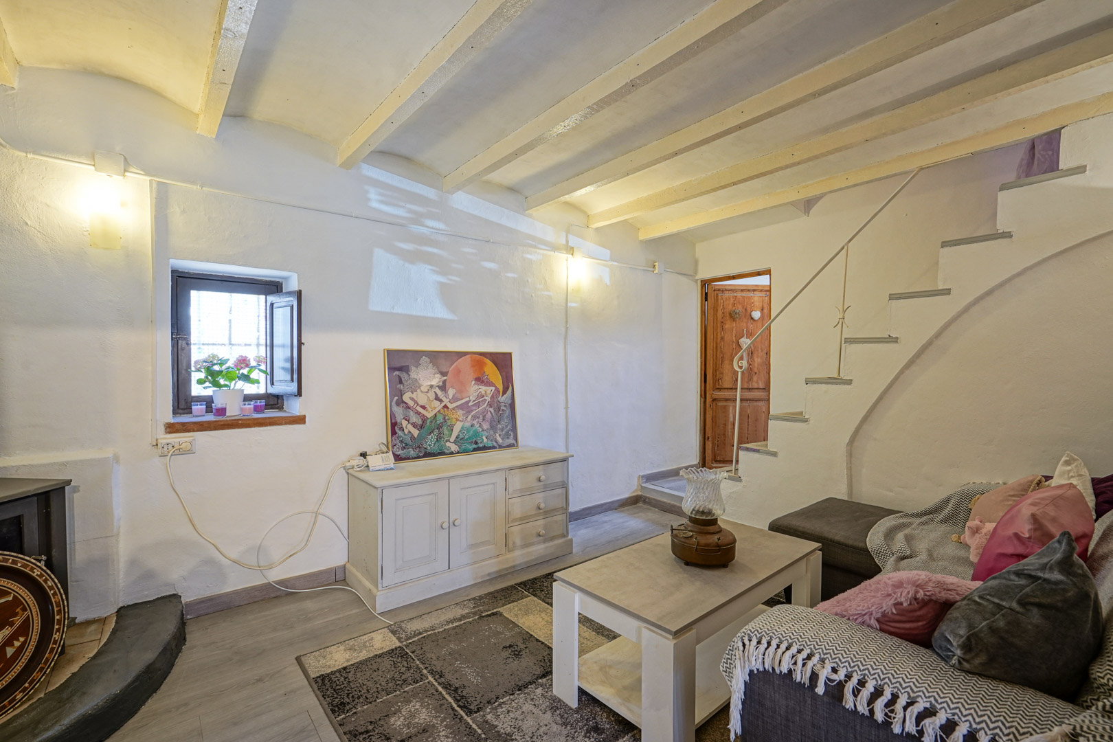 Segundo salón con vigas de madera a la vista y escalera blanca hacia la planta superior. Destacan los gruesos muros tradicionales y la luz natural de la ventana.